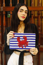 Load image into Gallery viewer, Woman holding a blue and white striped clutch with a red crab design against a wooden background.