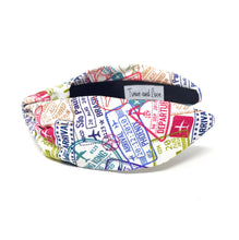 Load image into Gallery viewer, Colorful travel-inspired headband with a top knot design and multicolored passport stamp pattern on white fabric.