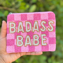 Load image into Gallery viewer, A hand holds a pink beaded pouch with a pink and light pink checkered pattern and the words 'BADASS BABE' in raised gold and white lettering, set against a blurred green outdoor background.