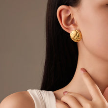 Load image into Gallery viewer, A close-up of a woman wearing a textured, spherical gold stud earring, with her hand resting near her neck against a neutral warm gray background.