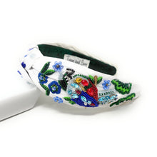 Load image into Gallery viewer, White hand-beaded knotted headband featuring a colorful jeweled bird, blue flowers, green leaves, and sparkling crystal embellishments. Handmade statement hair accessory by Twine and Love.