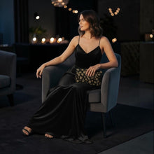 Load image into Gallery viewer, Woman in a black evening gown sitting on a chair in a dimly lit room with decorative lights.