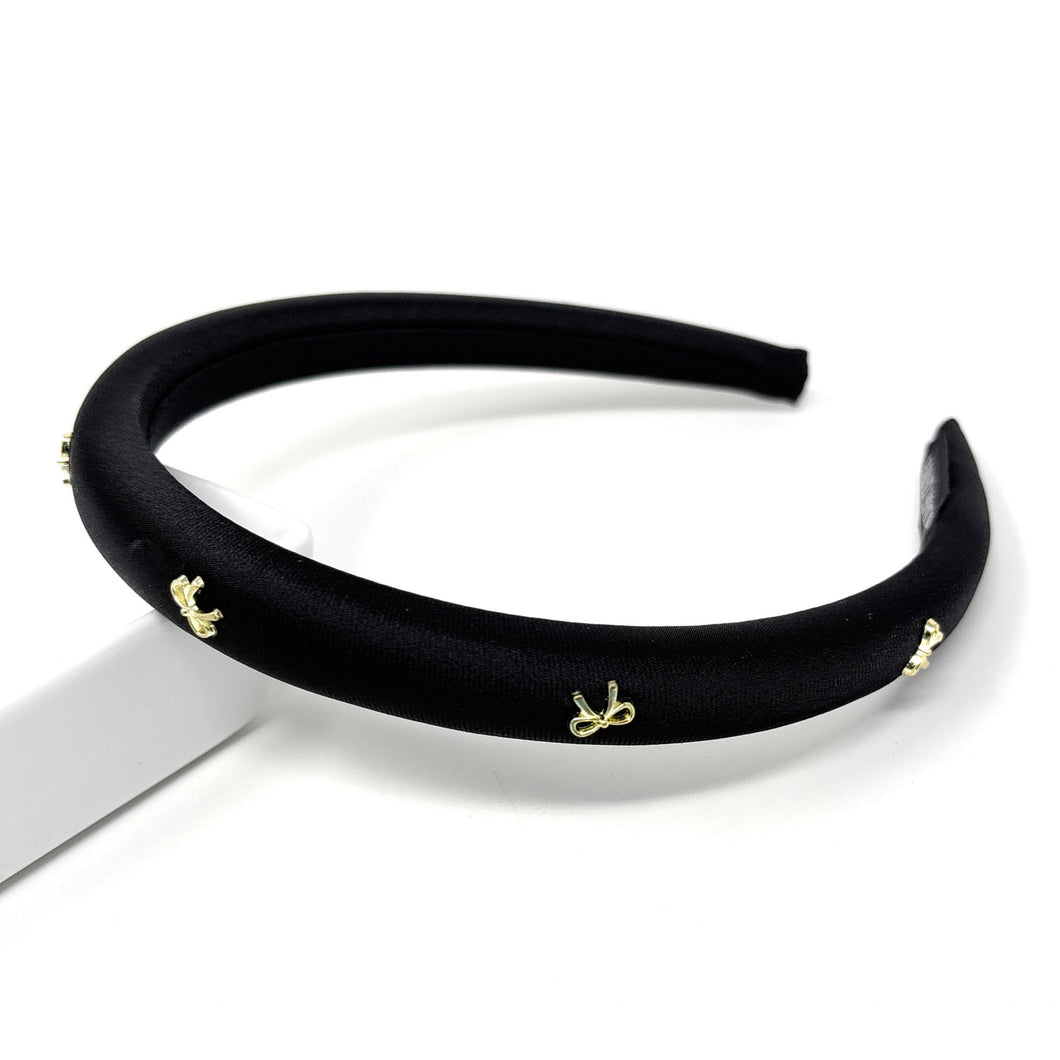 Elegant black satin headband adorned with small gold bow embellishments, displayed against a plain white background, conveying a stylish and chic tone.