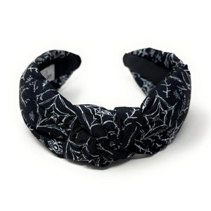 A black fabric headband with a knotted design is patterned with white spiderwebs and spiders. The mood is edgy and Halloween-themed.