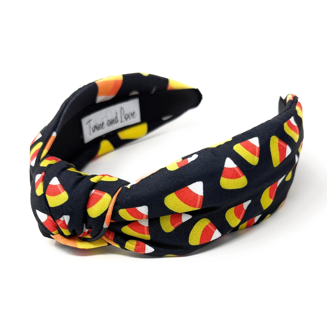 A black headband with colorful candy corn print, featuring white, orange, and yellow. Fun, Halloween-themed accessory.