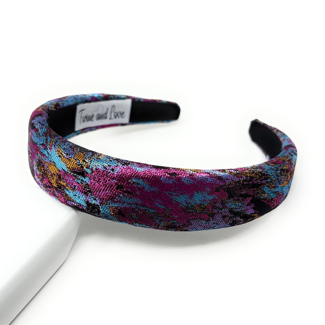 Elegant headband with a vibrant pattern of pink, blue, and gold accents on a textured fabric. It rests on a white surface, exuding a bold, stylish tone.