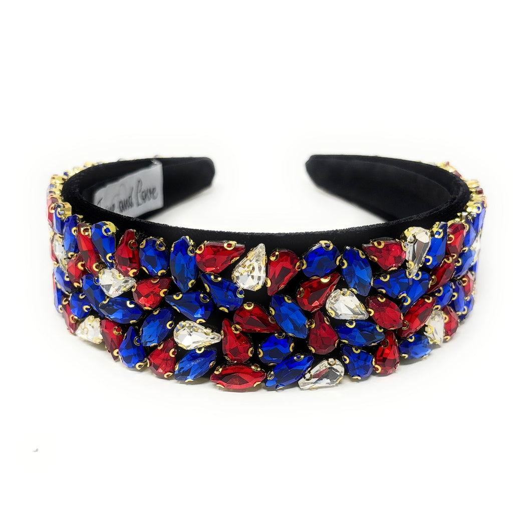 A wide, black headband adorned with vibrant red, blue, and clear gemstones arranged in clusters. The design conveys elegance and boldness.