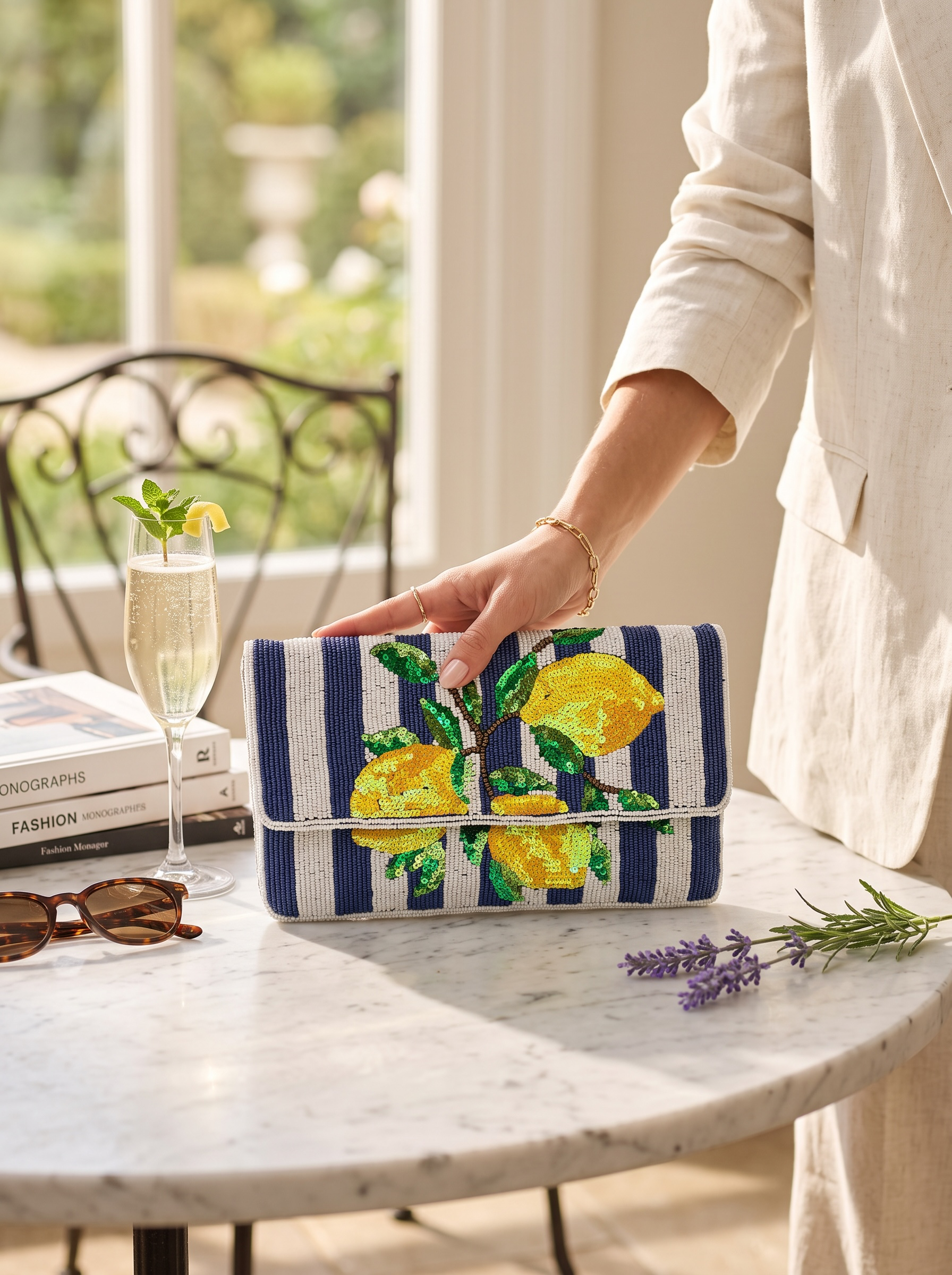 A navy and white striped clutch bag with a yellow lemon and green leaf floral pattern, held by a person wearing a cream linen blazer. The scene is set on a white marble table with a glass of champagne, books, sunglasses, and lavender sprigs in a bright, sunlit room.