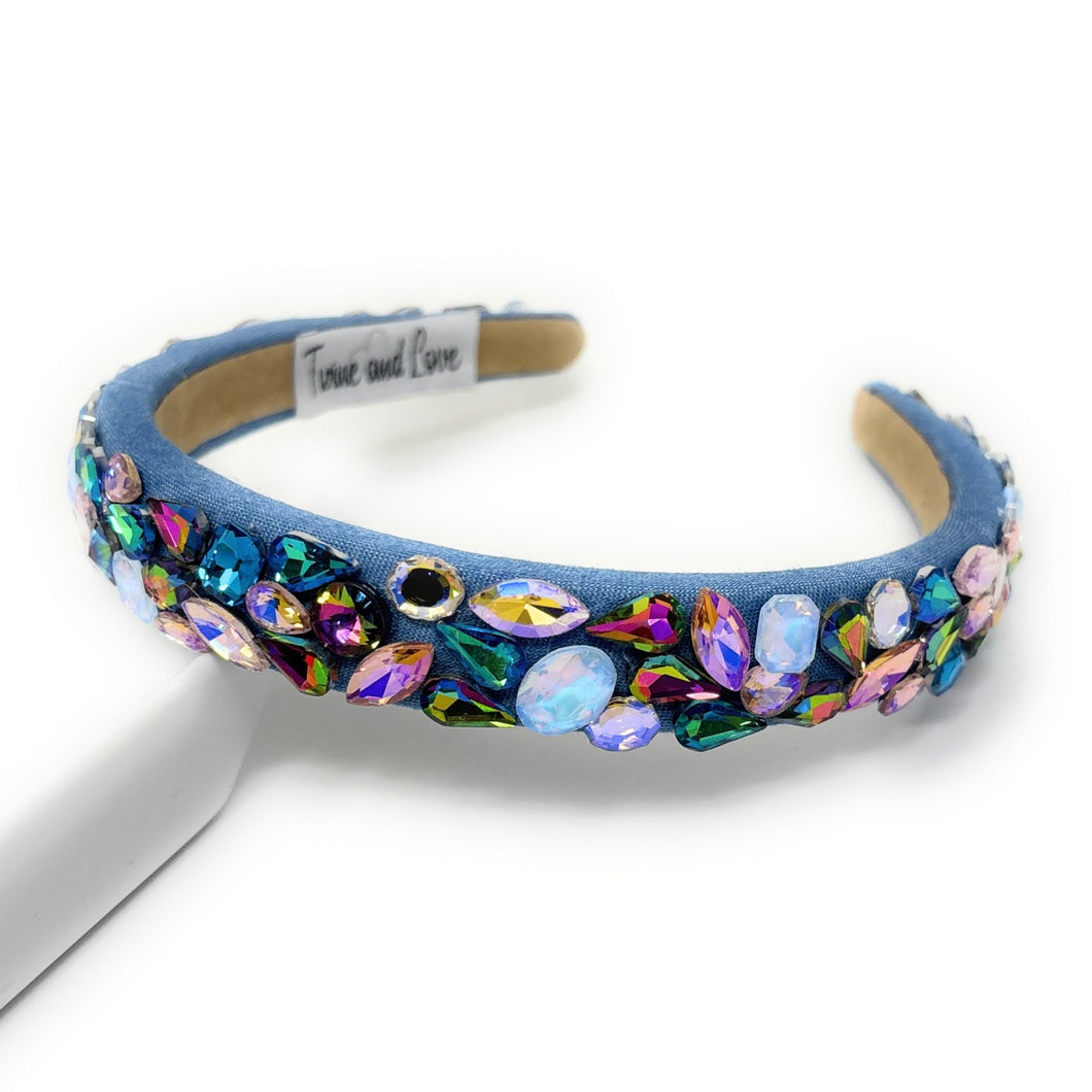 A vibrant, gemstone-encrusted headband with a blue fabric base adorned with multicolored jewels, including blue, purple, and gold, emanating a luxurious and playful vibe.