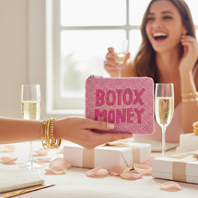 Load image into Gallery viewer, A pink textured zip pouch with the words 'BOTOX MONEY' in raised pink letters is held in a hand over a table with champagne glasses, gift boxes, and scattered rose petals. A smiling woman is blurred in the background near a window.