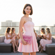 Load image into Gallery viewer, A woman stands on a rooftop terrace wearing a light pink strapless mini dress and holding a pink clutch that reads 'BOTOX MONEY'. Other women in similar pink outfits are seated in the blurred background.