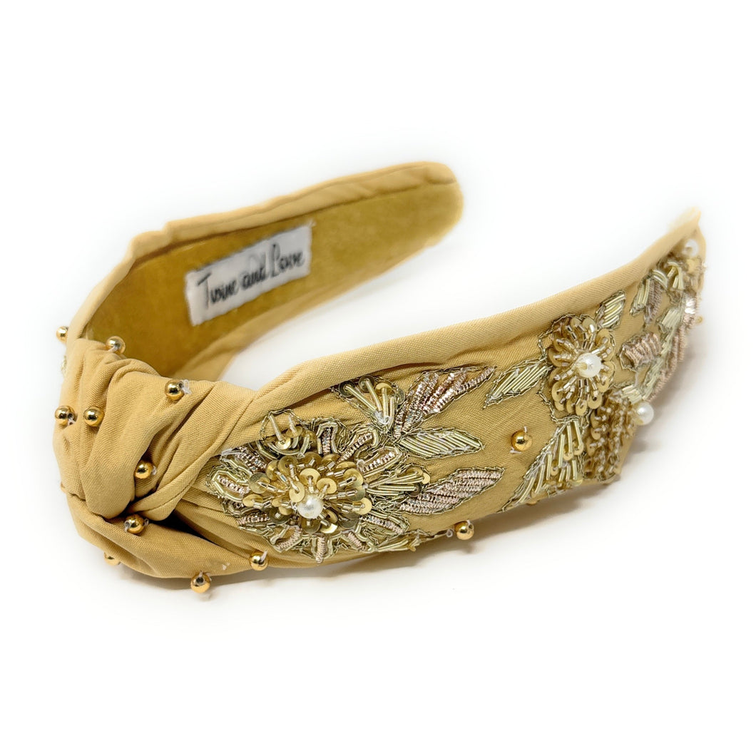 Golden padded headband adorned with intricate floral embroidery and pearl accents, exuding elegance and sophistication against a plain backdrop.