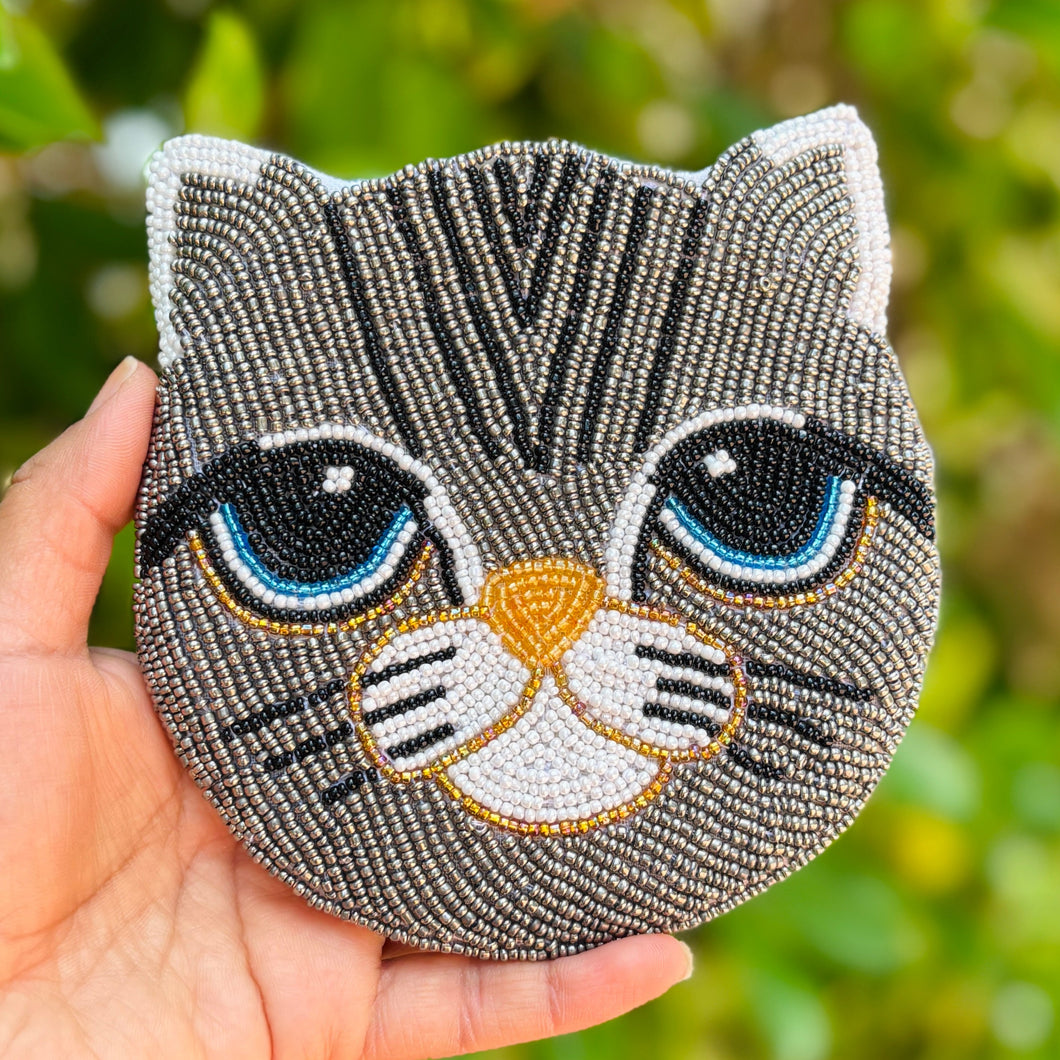 Beaded cat face with large blue eyes and a gray pattern. The intricate design features white ears and nose, and black whisker accents. Cute and whimsical.