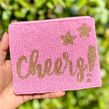 Load image into Gallery viewer, A pink beaded pouch with the word 'Cheers!' written in a golden script, accompanied by stars, held in a hand against a blurred greenery background.