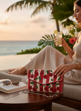 Load image into Gallery viewer, A woman in a beige dress sits by an ocean view at sunset, holding a cocktail while wearing a red and green cherry patterned clutch bag with a silver chain strap on a wooden table.