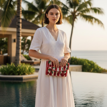 Load image into Gallery viewer, A woman wearing a white embroidered dress stands by a resort pool holding a red and white striped clutch bag with green palm leaf patterns, with palm trees and ocean in the background.