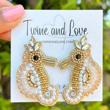Load image into Gallery viewer, Woman wearing a beige woven headband and gold seahorse statement earrings with a black one-shoulder top against a wooden backdrop.
