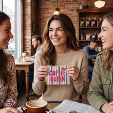 Load image into Gallery viewer, A woman in a beige sweater smiles while holding a colorful striped pouch that says 'COFFEE QUEEN' in pink letters, set in a cozy cafe with brick walls and other patrons in the background.