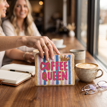 Load image into Gallery viewer, A woman holds a small woven clutch bag with pink 'COFFEE QUEEN' lettering and a gold crown on a striped multicolor background, set on a wooden cafe table with coffee cups, glasses, and a notebook nearby.