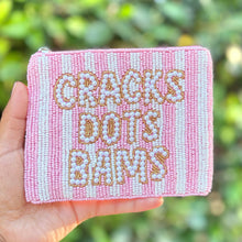 Load image into Gallery viewer, A hand holding a small pink and white beaded zip pouch with the text 'CRACKS DOTS BAMS' in gold-toned beads, set against a blurred green foliage background.