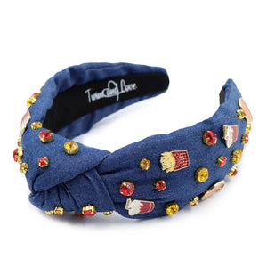 A blue denim headband with a knot design, decorated with red, gold, and clear rhinestones and small enamel fast-food-themed pins, including fries and sodas.