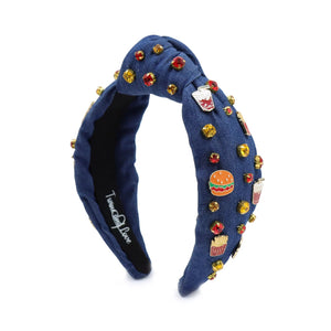 A blue denim headband with a knot design, decorated with red, gold, and clear rhinestones and small enamel fast-food-themed pins, including fries and sodas.