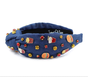 A blue denim headband with a knot design, decorated with red, gold, and clear rhinestones and small enamel fast-food-themed pins, including fries and sodas.