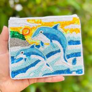 Beaded pouch featuring two blue dolphins jumping over stylized ocean waves. Background includes a yellow sun with spirals and green hills.
