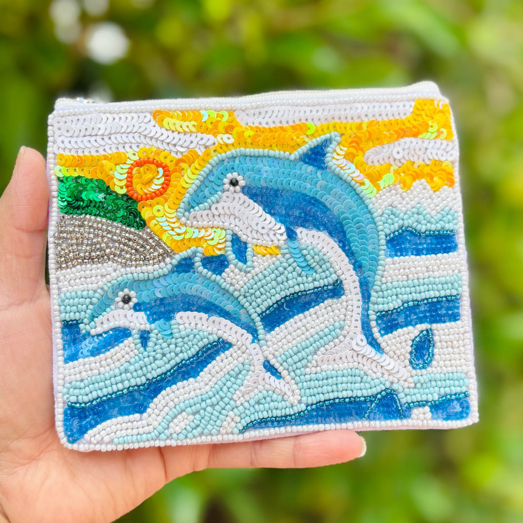Beaded pouch featuring two blue dolphins jumping over stylized ocean waves. Background includes a yellow sun with spirals and green hills.