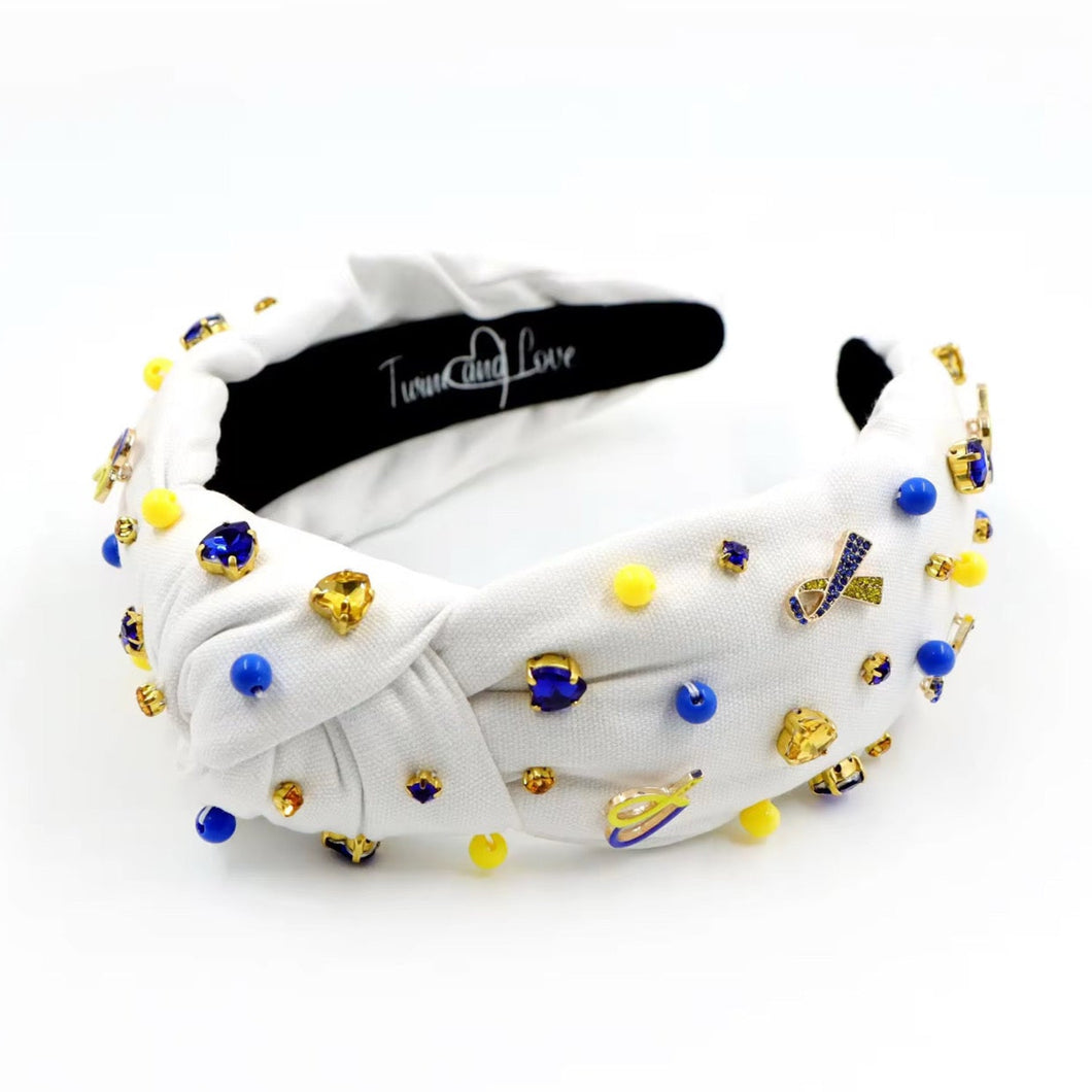 White padded headband with a twisted design, adorned with vibrant blue, yellow gemstones and down syndrome awareness charms. 