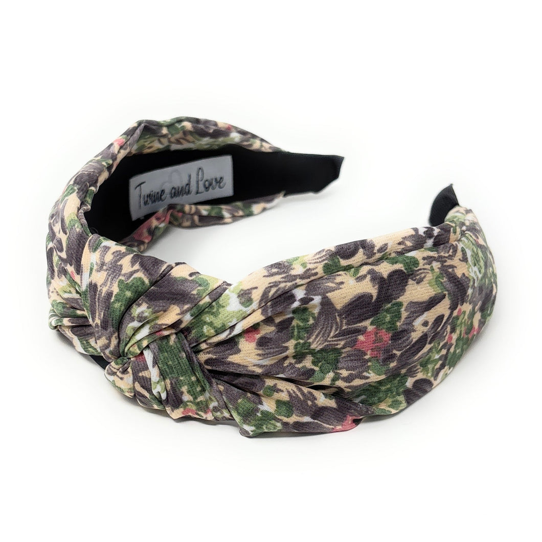 A floral patterned headband with a knot design, featuring shades of green, purple, and pink. It sits against a plain white background, conveying a stylish and feminine tone.