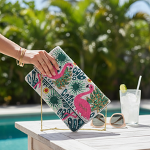 Load image into Gallery viewer, A hand holds a rectangular clutch bag with a tropical flamingo print, featuring pink flamingos, green palm leaves, and blue accents, set against a sunny outdoor poolside background with sunglasses and a drink on a table.