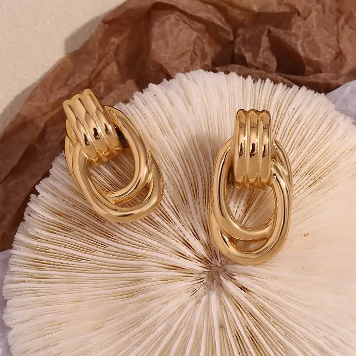 Gold-tone earrings with oval loops and ridged details, resembling linked chains. They convey a modern and elegant style, set against a plain white background.