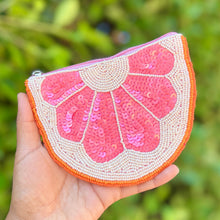 Load image into Gallery viewer, A hand holding a small semicircular sequined pouch designed to look like a pink grapefruit or citrus slice, with an orange border and cream-colored beadwork, set against a blurred green foliage background.