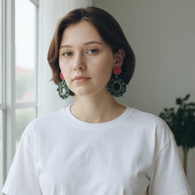 Load image into Gallery viewer, Woman wearing a white t-shirt and beige pants with green wreath earrings indoors.