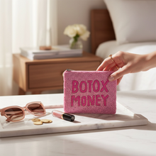 Load image into Gallery viewer, A pink textured zip pouch with the words 'BOTOX MONEY' in raised pink letters sits on a marble tray beside sunglasses, coins, and lipstick. A hand holds the pouch in a softly lit bedroom setting.