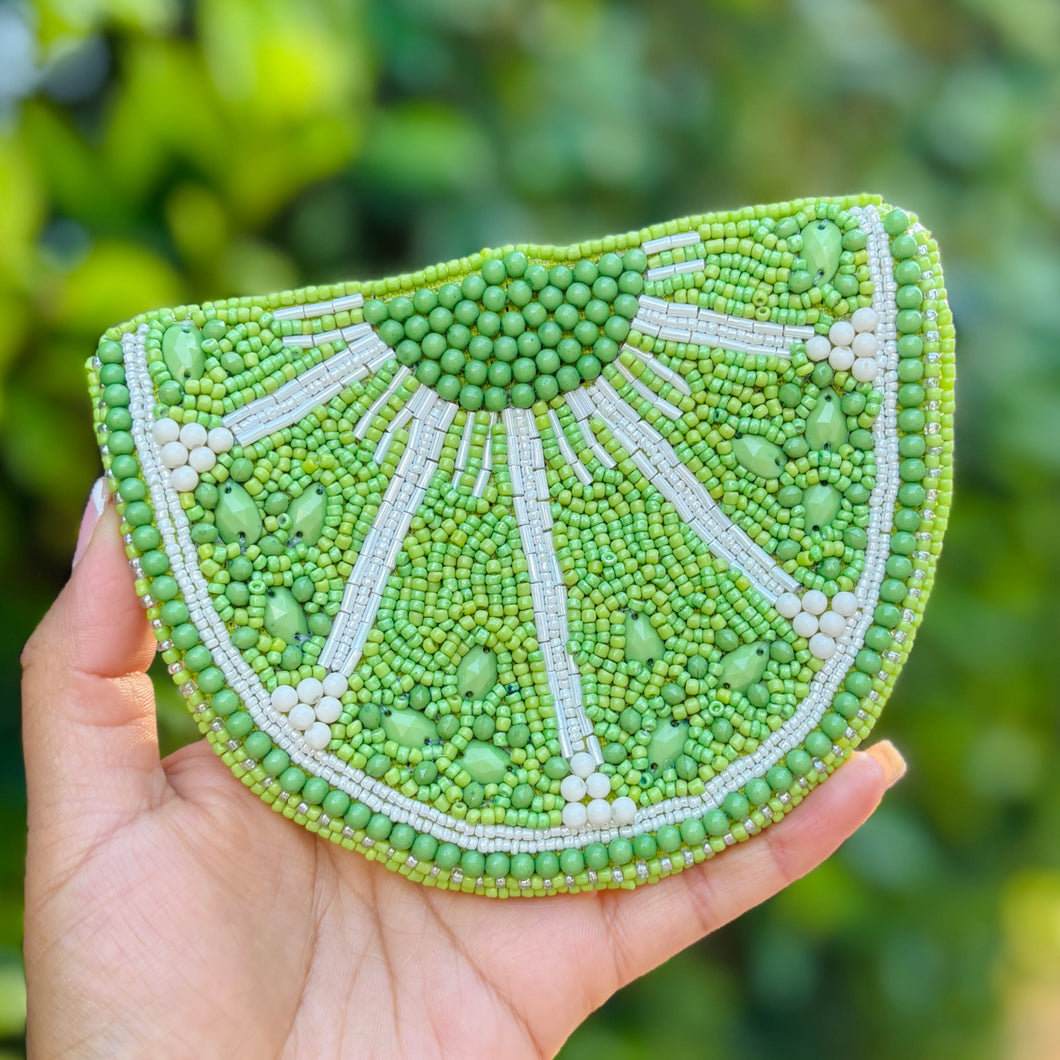 A beaded pouch shaped like a lime slice, featuring vibrant green and white beads. The design is textured and playful, evoking a fresh, summery feel.