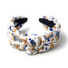 Load image into Gallery viewer, A knotted fabric headband featuring a white background with a festive pattern of gold and navy blue menorahs, stars, and stripes, shown on a clean white background.
