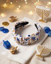 Load image into Gallery viewer, A knotted fabric headband with a white background and blue and gold Hanukkah-themed patterns, displayed on a white napkin with festive holiday decorations including dreidels, candles, and string lights.