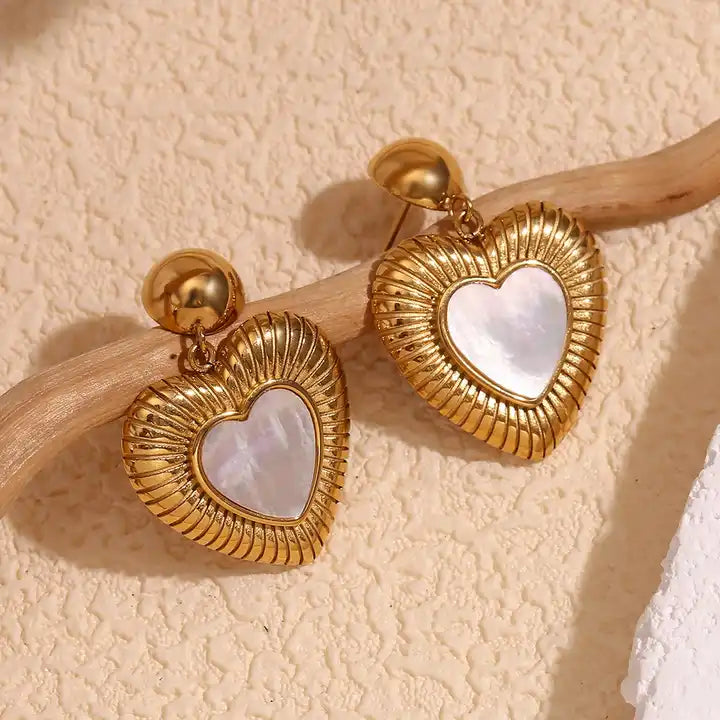 Gold heart-shaped earrings with a ribbed texture and a central mother-of-pearl heart. They hang from small gold spheres, evoking elegance and romance.