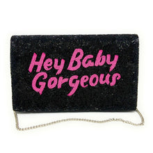 Load image into Gallery viewer, Woman holding a black hand-beaded clutch featuring bright pink “Hey Baby Gorgeous” lettering against a light blue wall.