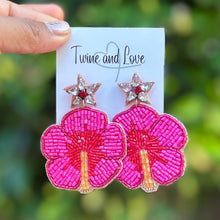 Load image into Gallery viewer, A hand holds a small white card displaying a pair of bright pink flower-shaped earrings with star studs, against a blurred green outdoor background. The card is branded 'Twine and Love'.