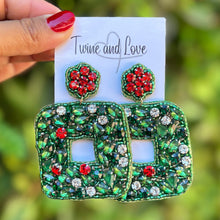Load image into Gallery viewer, Hand holding a card with green and red embellished earrings against a blurred green background