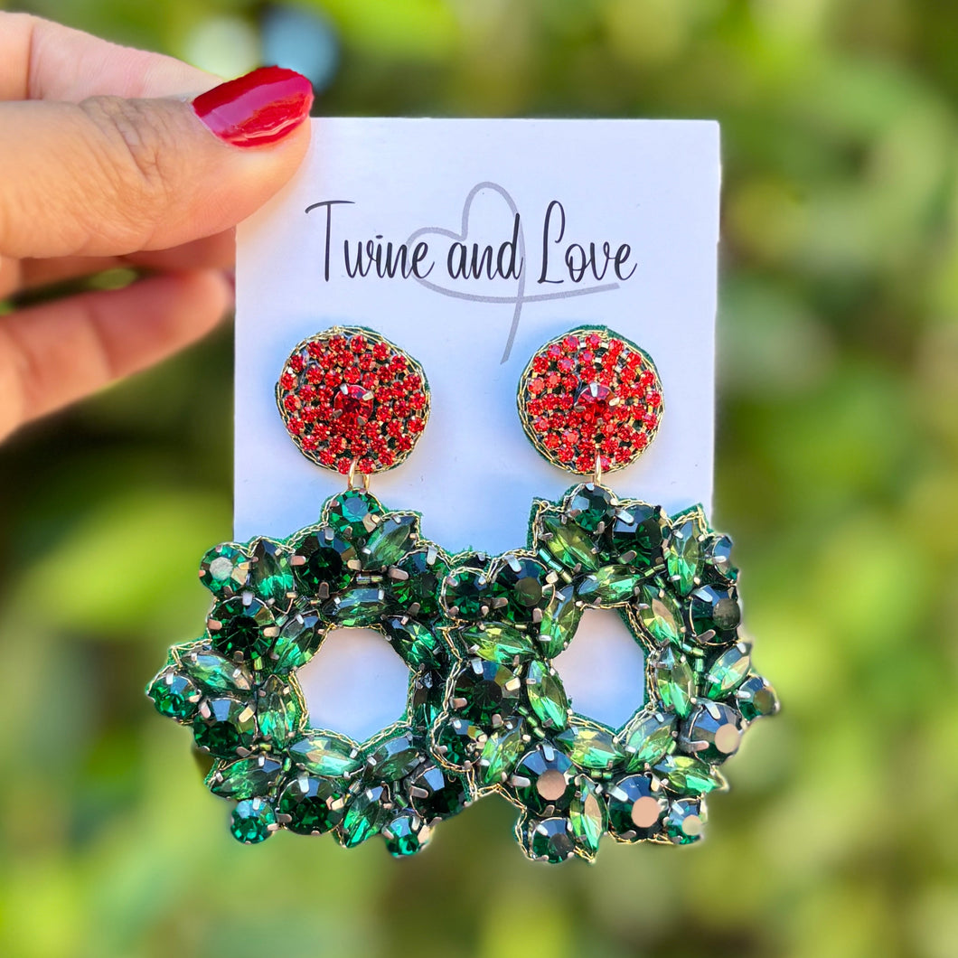 Hand holding a card with red and green wreath earrings against a blurred green background