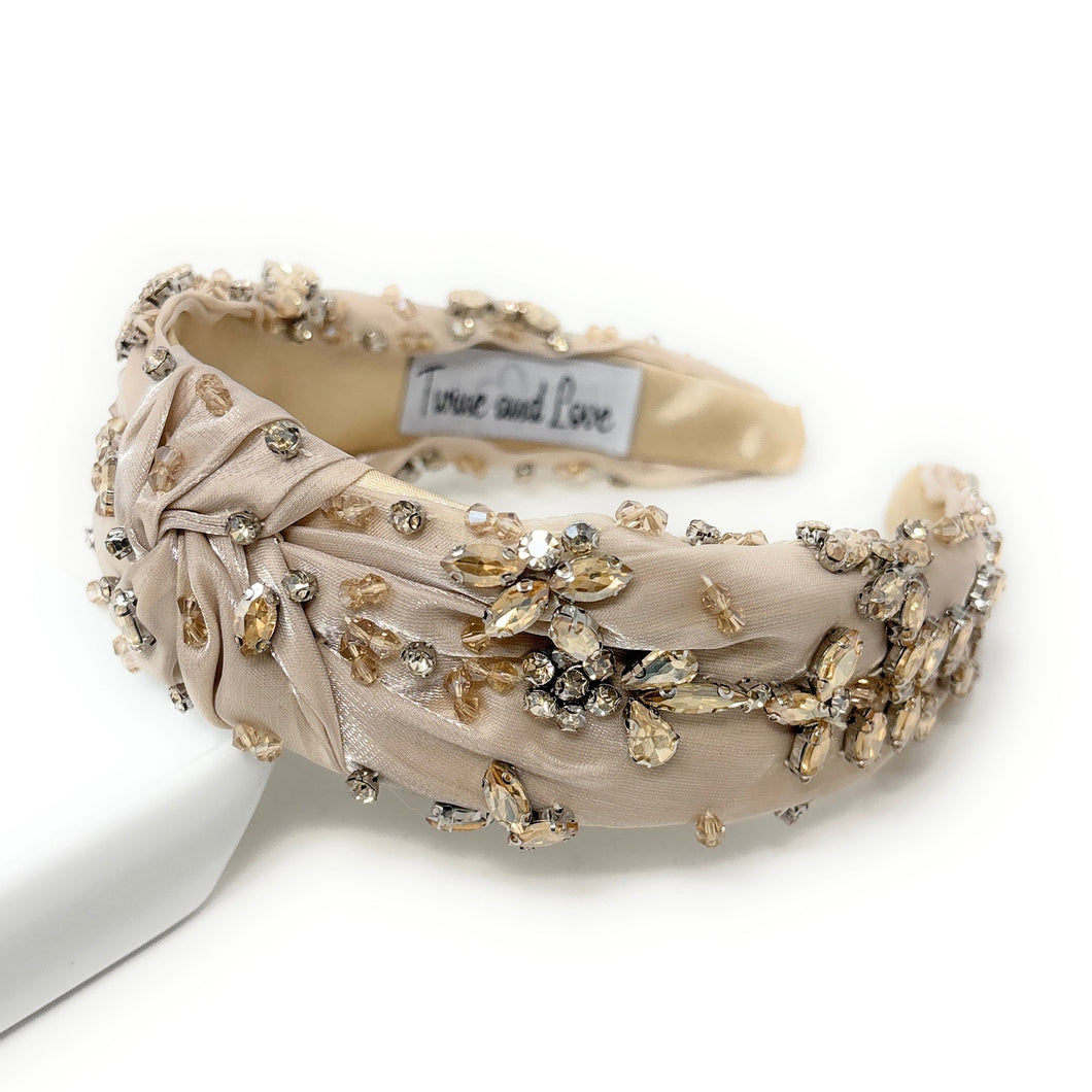 Elegant beige headband adorned with sparkling crystals and gemstones. The luxurious fabric is delicately ruched, creating a sophisticated and glamorous look.