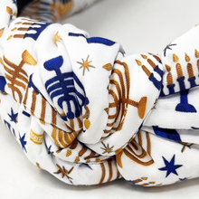 Load image into Gallery viewer, A close-up of a knotted fabric headband or hair accessory featuring a white base with blue and gold holiday-themed patterns, including menorahs and stars