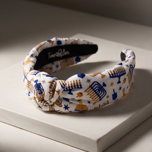 Load image into Gallery viewer, A knotted fabric headband with a white background and a colorful pattern of blue and gold holiday symbols, displayed on a white block with warm directional lighting.