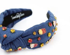 A blue denim headband with a knot design, decorated with red, gold, and clear rhinestones and small enamel fast-food-themed pins, including fries and sodas.
