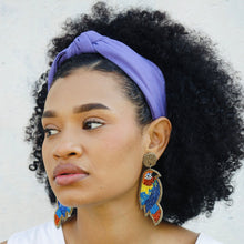Load image into Gallery viewer, Woman wearing a lavender knotted headband and colorful beaded parrot earrings, styled with a white sleeveless top and natural curly hair.