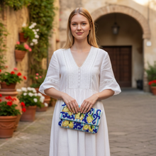 Load image into Gallery viewer, A woman stands in a sunlit courtyard wearing a white dress and holding a blue, yellow, and green patterned clutch bag featuring lemons on several branches.